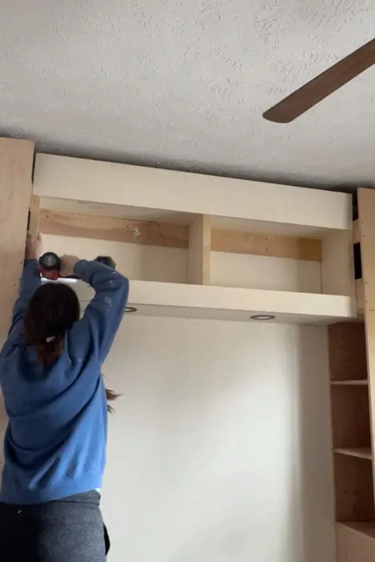 A DIYer attaching a face frame to a built-in headboard with storage, using a power drill. The structure is starting to take on a finished look, with recessed lighting installed in the lower section.
